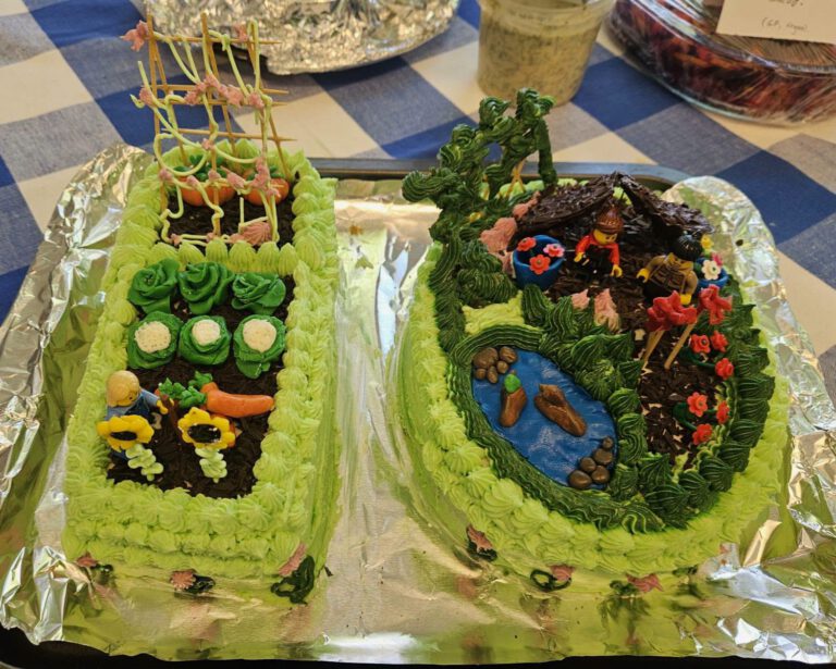Close up of two mainly green decorated cakes with intricate icing depicting vegetables, people gardening, a duck pond and two lego figures. The cakes are shaped as the figure 1 and 0. All on a blue and white gingham tablecloth.