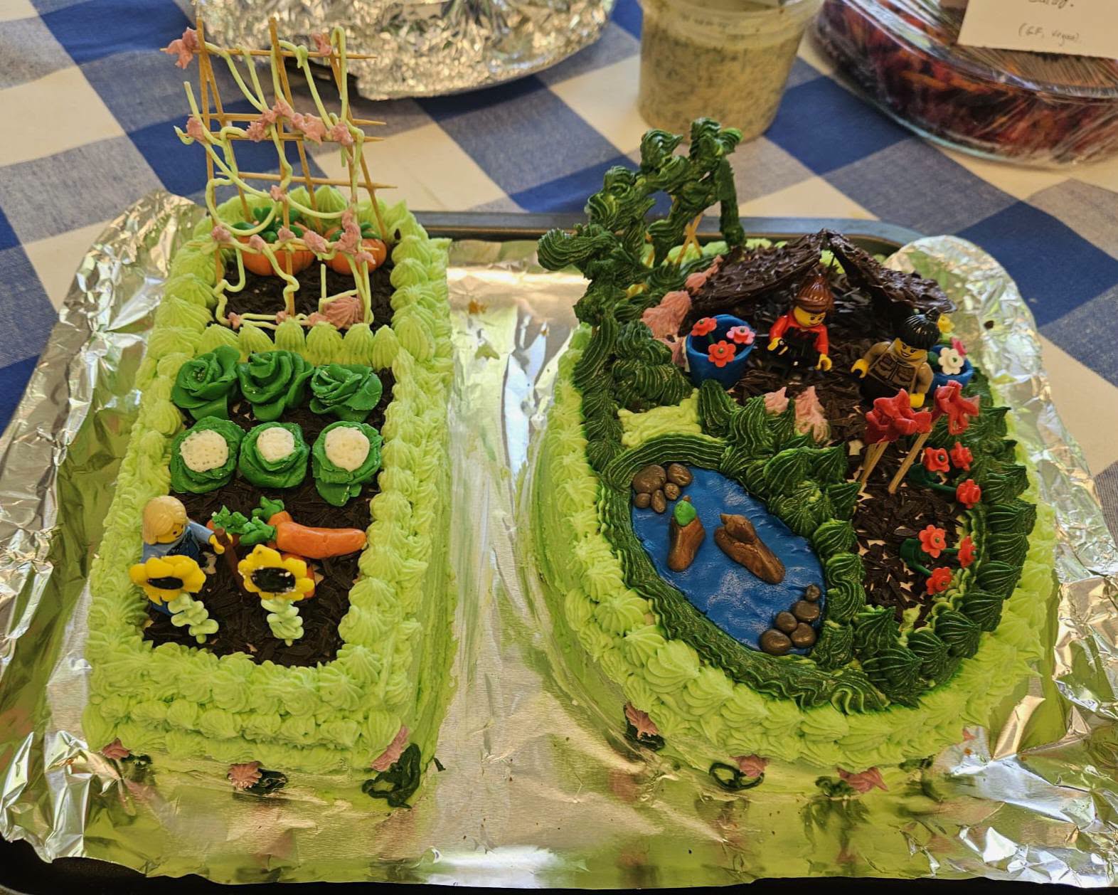 Close up of two mainly green decorated cakes with intricate icing depicting vegetables, people gardening, a duck pond and two lego figures. The cakes are shaped as the figure 1 and 0. All on a blue and white gingham tablecloth.