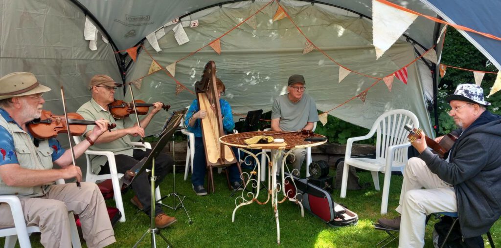 Musicians under an awning, on white chairs, playing music. Two fiddles, one guitar and a small harp.