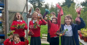A group of seven girls standing holding various bags and trays of freshly-picked vegetables. They are standing beside a large polytunnel with potted plants to one side and a hedge in the background.