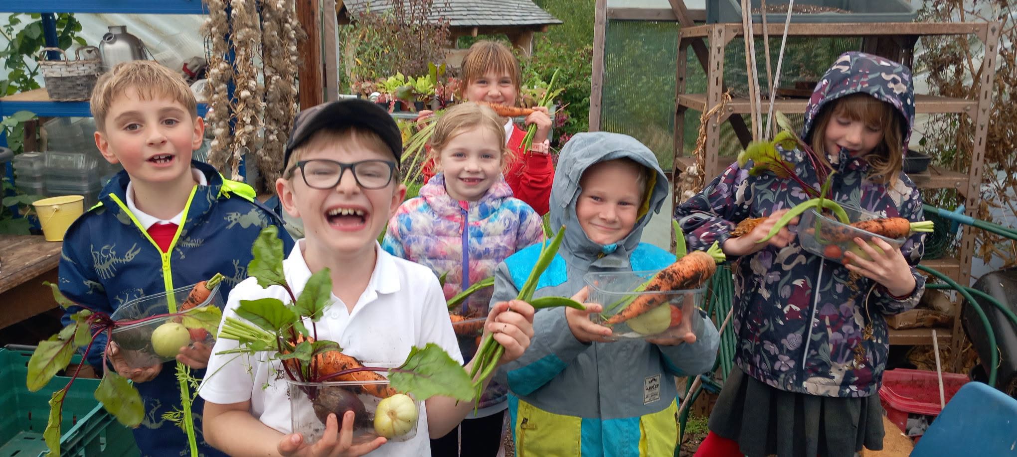 Group of six children in a polytunnel, each smiling towards the camera and holding various freshly-picked vegetables