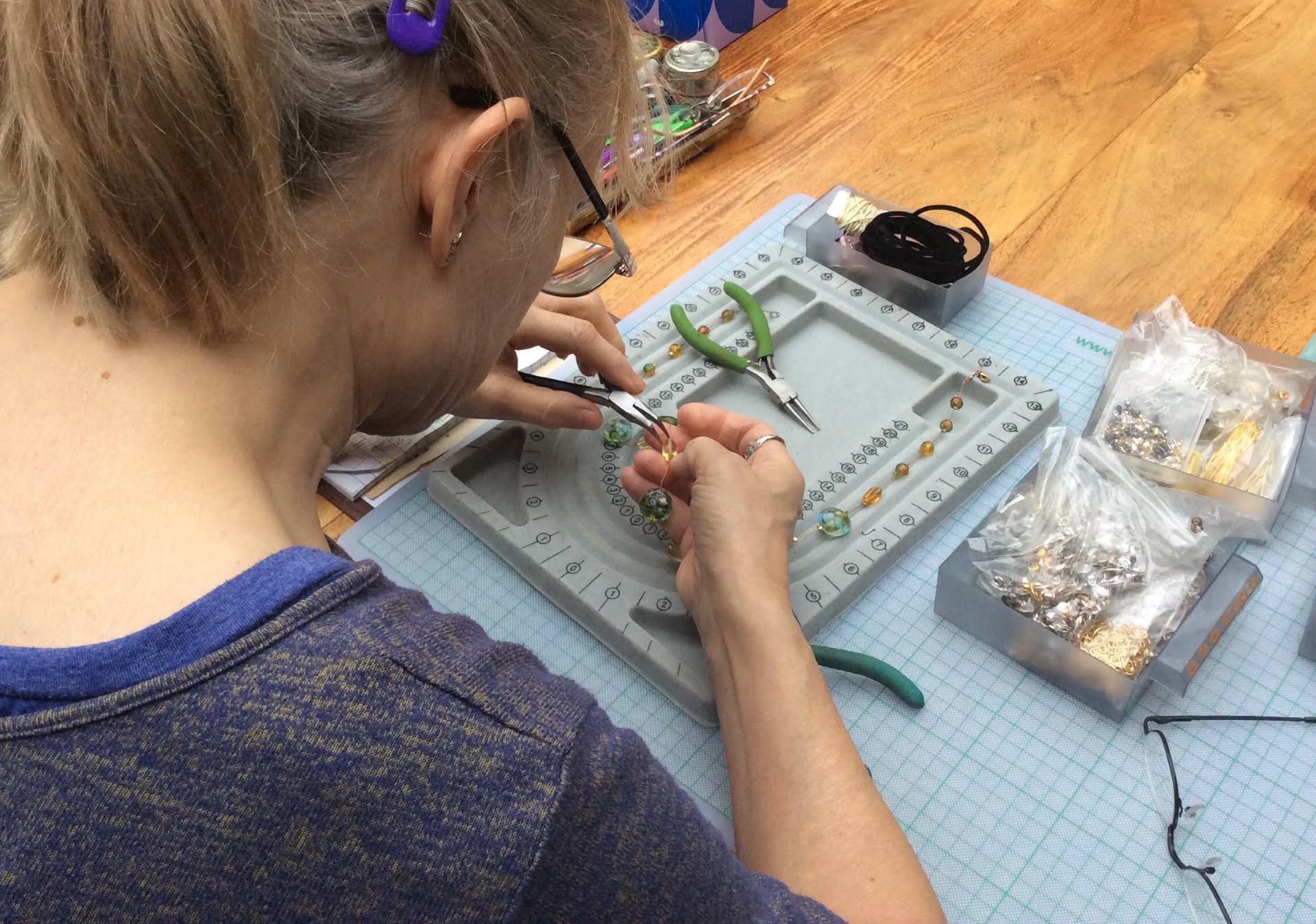 Seen from above, a woman is repairing some jewellery with small pliers. Various tools and accessories are on the desk in front of her.